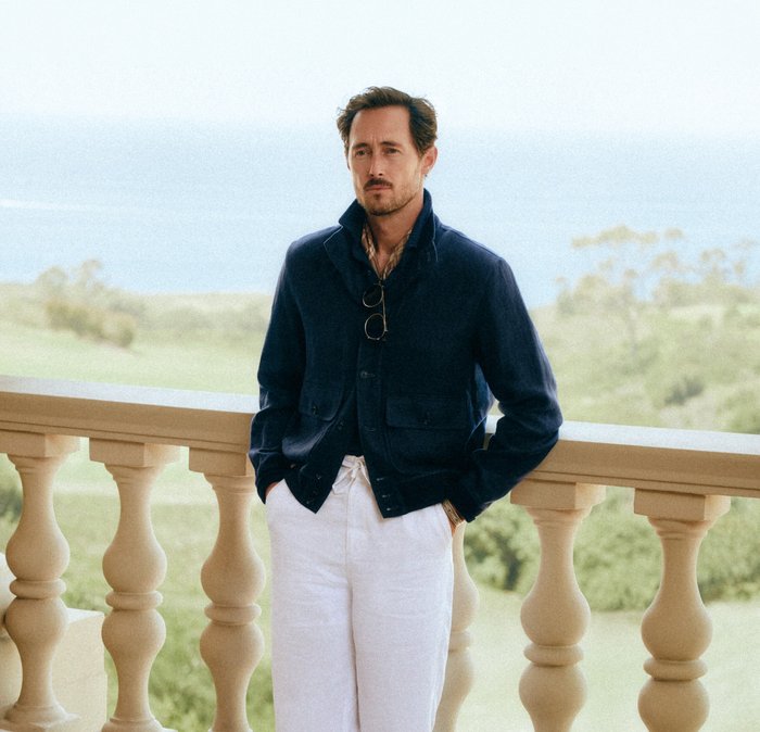 Uomo con giacca blu navy e pantaloni bianchi in piedi con le mani nelle tasche su un balcone in pietra con vista su un paesaggio verde.