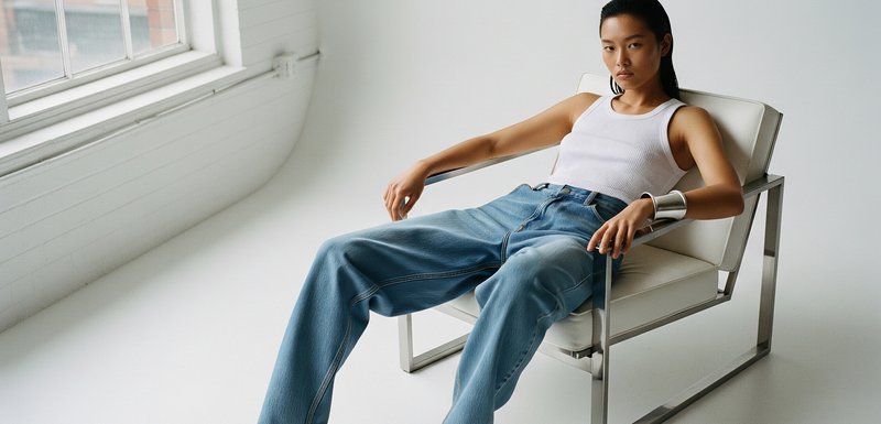 Jean en denim bleu clair taille haute avec des jambes larges, associé à un débardeur blanc ajusté, assis dans une chaise moderne avec un cadre en argent.