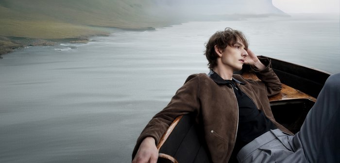 Veste en daim marron avec un col noir, superposée à une chemise noire et un pantalon gris. Le décor présente des eaux calmes et des couleurs douces et atténuées.