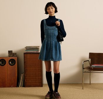 Sukienka ogrodniczka z denimu z plisowanym dołem w kolorze niebieskim. Modelka nosi granatowy golf i czarne skarpetki za kolano, uzupełnione brązowymi butami.