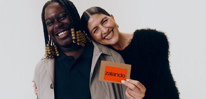 Zwei Freunde lächeln, einer hält eine Zalando-Geschenkkarte vor einem schlichten weißen Hintergrund.