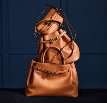 Drie tankleurige leren handtassen met gouden gespen gestapeld tegen een donkerblauwe wand met panelen.