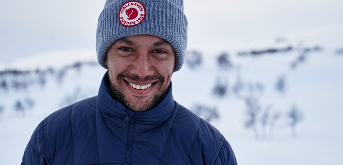Gepolsterte dunkelblaue Jacke mit hohem Kragen und Reißverschluss, graue gestrickte Mütze mit Logo. Lächelnde Person vor einer verschneiten Landschaft.