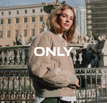 Jonge vrouw in een beige doorgestikte jas staat bij een decoratieve stenen fontein en een ijzeren hekwerk op een stedelijk plein. Het woord "ONLY" is over het beeld gelegd.