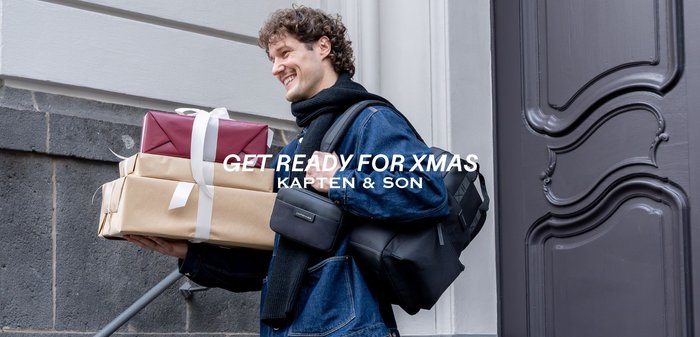 Černé a šedé tašky s hladkými texturami, jedna držená pod paží, tři dárkově zabalené krabice naskládané v červeném, hnědém a bílém papíru.