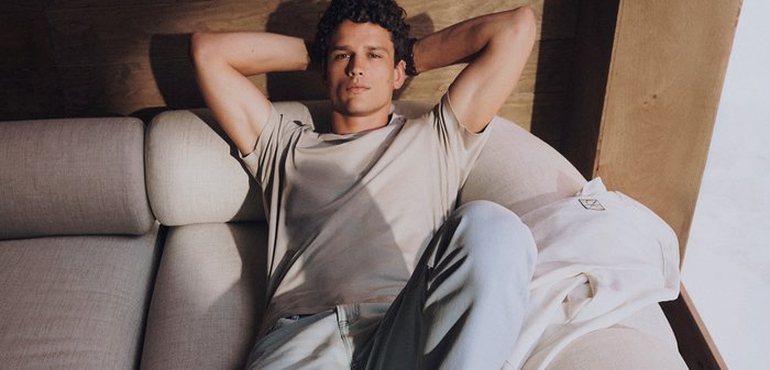 En ung man i vardagskläder lutar sig bakåt på en beige soffa med armarna bakom huvudet, i ett mjukt upplyst rum med en bakgrund av träpaneler.