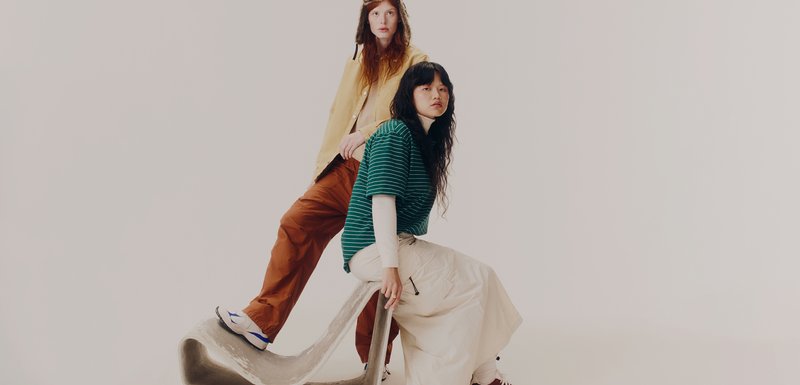 Deux modèles posent sur une structure grise texturée. L'un porte une veste beige et un pantalon marron ; l'autre a une chemise rayée verte et un pantalon crème.