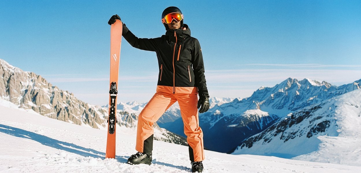 Suusavarustus sisaldab paari oranže suuski mustade kinnitustega. Suusataja kannab musta jope ja oranže pükse, taustal lumevalge mägi.