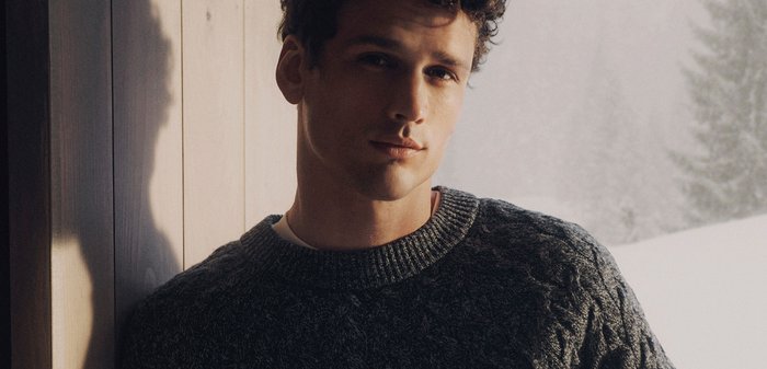 Ung man med lockigt hår som bär en mörk kabelstickad tröja, lutande mot en trävägg med en snötäckt skog synlig utanför.