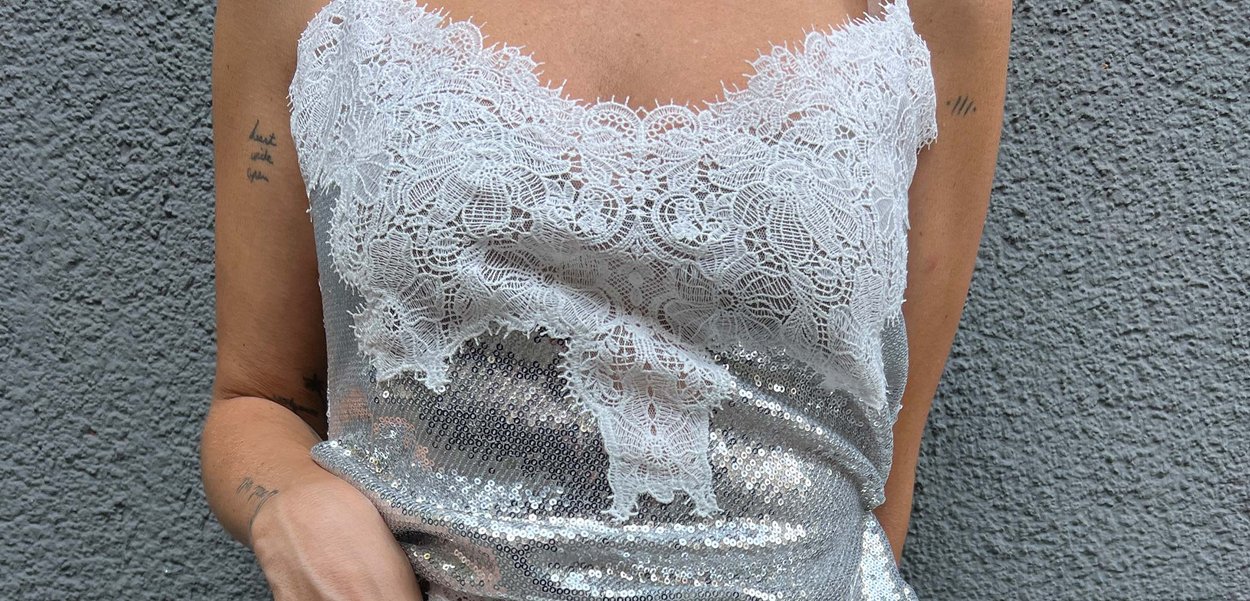 Camiseta de tirantes plateada con lentejuelas y un encaje blanco en el escote. Presenta tirantes finos y un diseño ajustado. Textura suave en general.