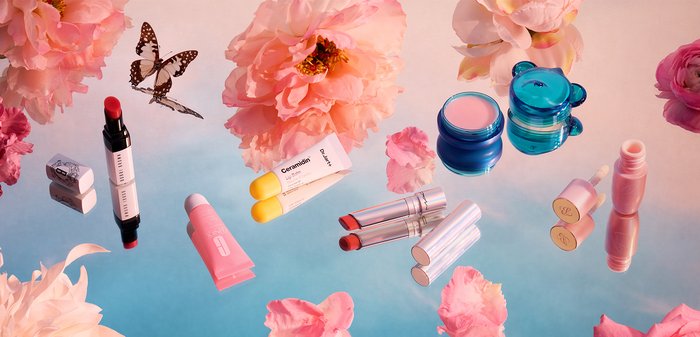 Divers cosmétiques sur une surface réfléchissante, présentant des tubes colorés, des rouges à lèvres en stick et des pots, avec des fleurs roses et un papillon à proximité.