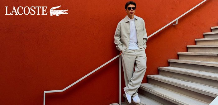 Jeune homme en veste et pantalon beige debout sur des escaliers contre un mur rouge, portant des lunettes de soleil et des baskets blanches, avec un logo Lacoste en superposition.