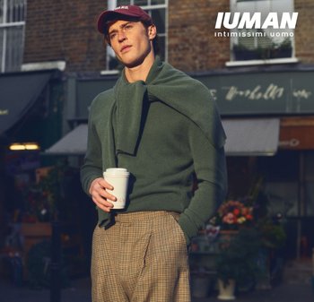 Maglione verde con una sciarpa drappeggiata sulle spalle, pantaloni color cammello a quadri e una tazza di caffè bianca in mano. Il look è completato da un berretto da baseball rosso.