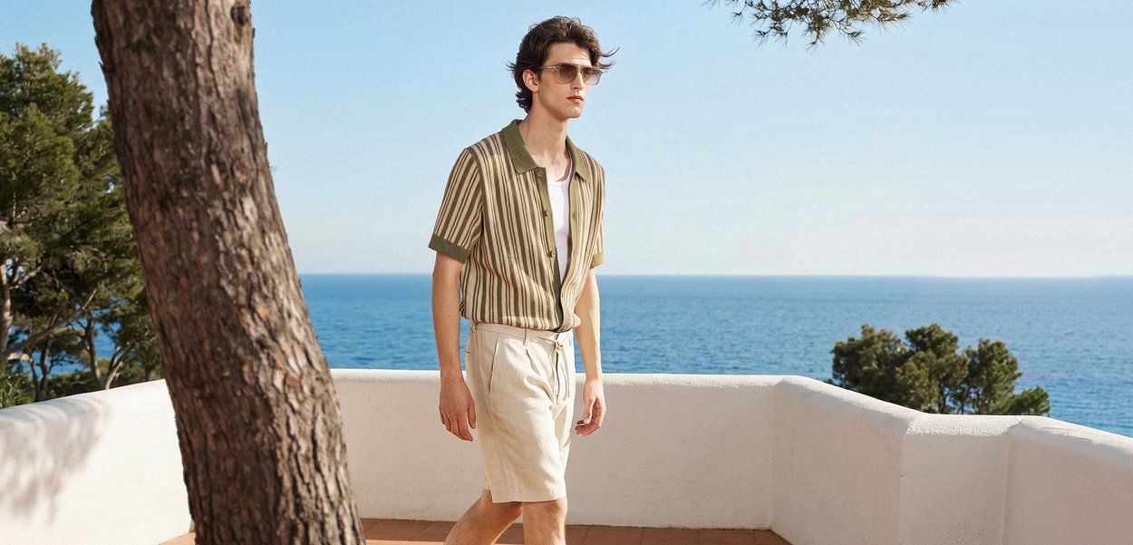 Junger Mann mit Sonnenbrille, gestreiftem kurzärmligem Hemd und hellen Shorts läuft auf einer Terrasse mit Blick aufs Meer.