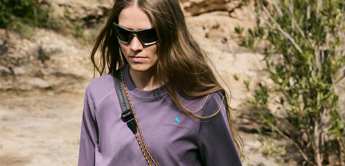 Jonge vrouw met zonnebril en een paarse lange mouwen blouse buiten, met rotsachtig terrein en groene struiken op de achtergrond.