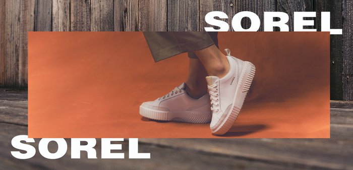 White Sorel branded sneakers worn by a person standing on an orange surface with wooden floor and planks background.