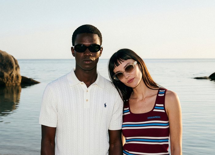 Άνδρας με λευκό πόλο και γυαλιά ηλίου στέκεται δίπλα σε γυναίκα με ριγέ φανελάκι και γυαλιά ηλίου δίπλα σε ήρεμη θάλασσα με βράχια στο φόντο.