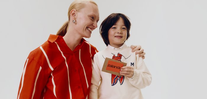 Volwassene met een rood gestreept hemd die glimlacht naar een kind dat een Zalando-cadeaubon vasthoudt, beiden staan tegen een effen witte achtergrond.