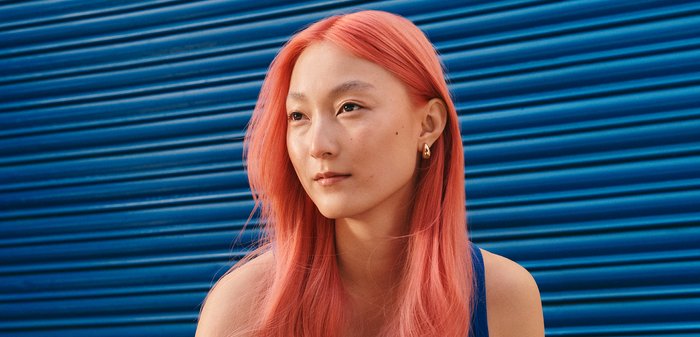 Junge Frau mit langen pinkfarbenen Haaren und blauem ärmellosen Oberteil, die vor einer blauen, wellenförmigen Metallwand sitzt und nachdenklich geradeaus schaut.
