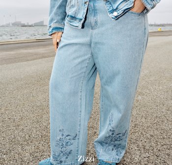 Person iført lyseblå broderede vidde jeans og denimjakke, stående på et befærdet område nær vandet.