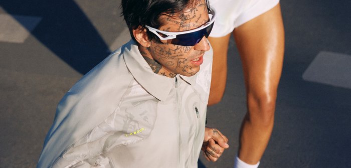 Homme avec des tatouages faciaux portant des lunettes de soleil de sport blanches et une veste claire, courant à côté d'une personne en short blanc sur une route ensoleillée.