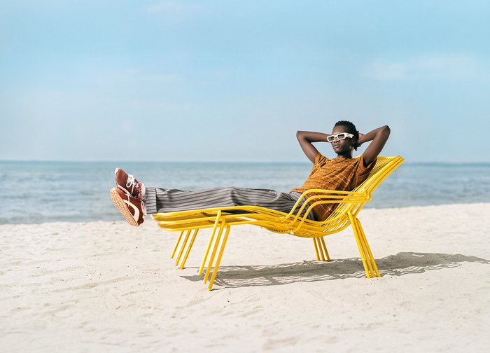 Personne portant des lunettes de soleil et des vêtements décontractés, allongée sur une chaise longue jaune sur une plage de sable au bord de la mer sous un ciel clair.
