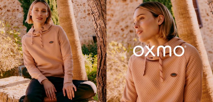 Junge Frau trägt einen pfirsichfarbenen Hoodie und schwarze Hose, sitzt im Schneidersitz auf einer Steinmauer im Freien, in der Nähe von Bäumen und Sträuchern.