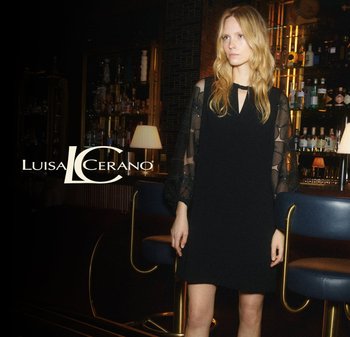 Black dress with sheer, patterned sleeves and a keyhole neckline. Smooth fabric, relaxed fit, paired with a modern bar setting in the background.