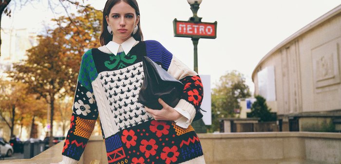 Jeune femme portant un pull coloré à motifs et une chemise avec col blanc s'appuie sur la rampe près d'une entrée du métro parisien, tenant un grand sac à main noir.