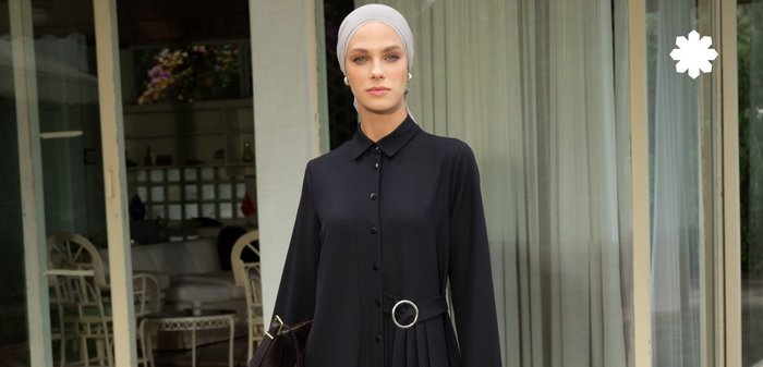 Robe longue noire avec une fermeture à boutons, une partie inférieure plissée et une ceinture avec une boucle circulaire en argent. Accompagnée d'un sac à main sombre. Tissu lisse.