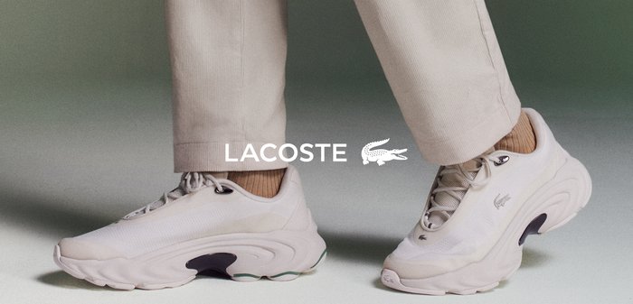 Baskets Lacoste, beige clair, avec une tige texturée, un bout arrondi et un design de semelle distinctif. Associées à un pantalon beige clair, sur un fond dégradé doux.