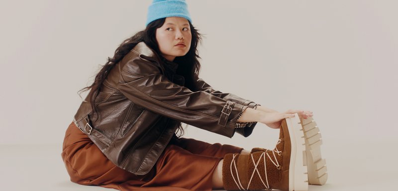 Femme en veste en cuir marron, jupe rouille et bonnet bleu assise et s'étirant, touchant ses bottes d'hiver beiges avec des semelles blanches.