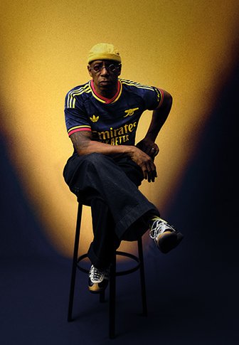Mann mit gelber Kappe, Brille und dunkelblauem Sporttrikot sitzt mit verschränkten Beinen auf einem Hocker vor einem gelben und dunklen Hintergrund.