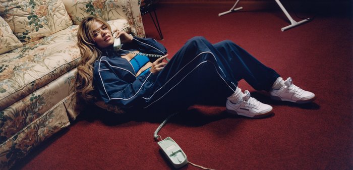 Mujer con chándal azul tumbada en un suelo de alfombra roja, hablando por un teléfono con cable junto a un sofá floral en una habitación poco iluminada.