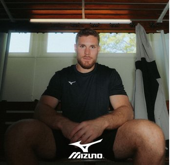 Camiseta atlética negra de manga corta, en postura sentada, con barba y pantalones cortos. Luz natural de las ventanas, banco de madera, logo de Mizuno visible.