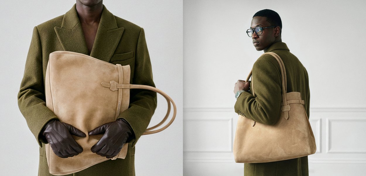 Homem com casaco verde-oliva e luvas pretas segurando e usando uma grande mala de mão bege em couro numa sala branca minimalista.