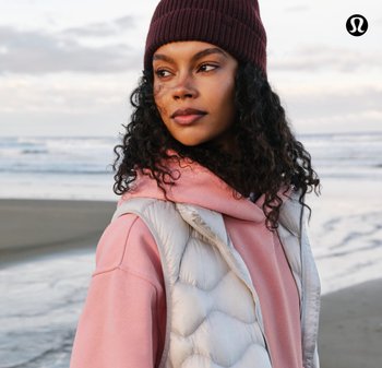 Lichtgewicht, donsachtige witte bodywarmer over een roze hoodie, gecombineerd met een donkere muts. Krullend haar valt over de schouders, met een strandachtergrond.
