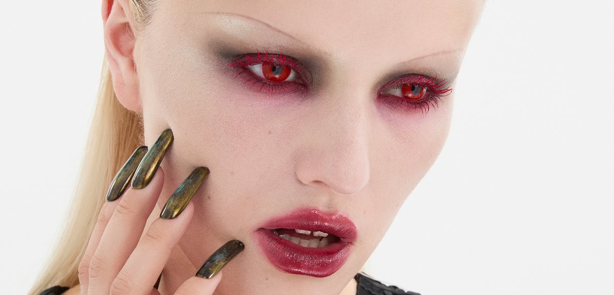 Nahaufnahme eines Gesichts mit heller Haut, kühnen roten Augen, dunklem Augen-Make-up und langen, metallisch grünen Nägeln sowie glänzend roten Lippen.