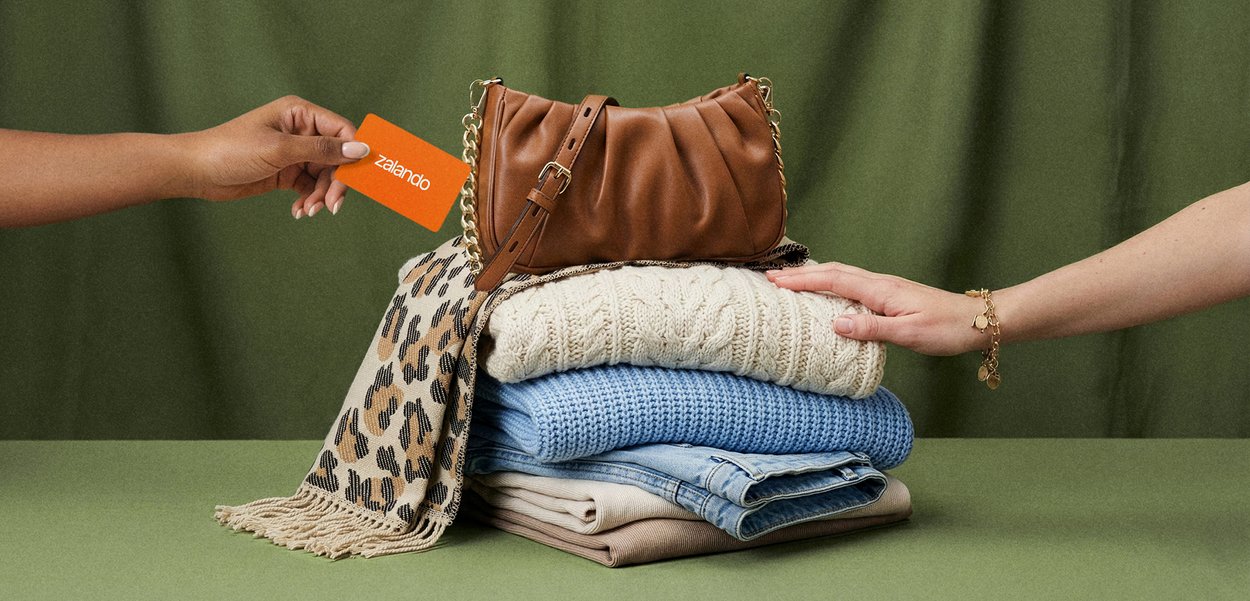 Persoon overhandigt een oranje Zalando-kaart, een andere reikt naar een bruine handtas op opgevouwen kleren en een sjaal met luipaardprint.