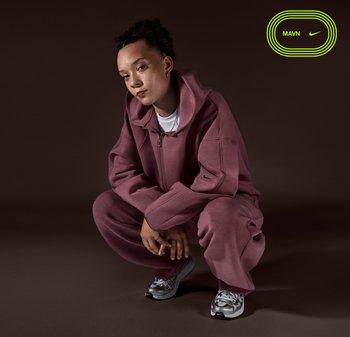 Person with curly hair squatting, wearing oversized mauve hoodie and pants with silver and white Nike sneakers on dark brown background.