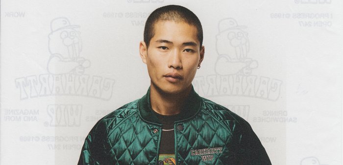 Chaqueta bomber acolchada verde con cuello acanalado, logo bordado "Carhartt WIP", usada sobre una camiseta gráfica negra que presenta un diseño minimalista.