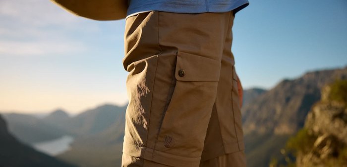 Person wearing tan cargo pants standing on a sunny mountain trail with distant peaks and valley in the background.