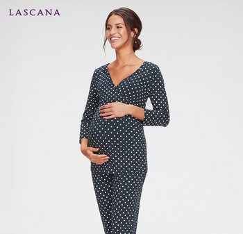 Schwangere Frau lächelt, trägt ein marineblaues langärmliges Oberteil mit Punkten und passende Hose, hält ihren Bauch sanft in den Händen.