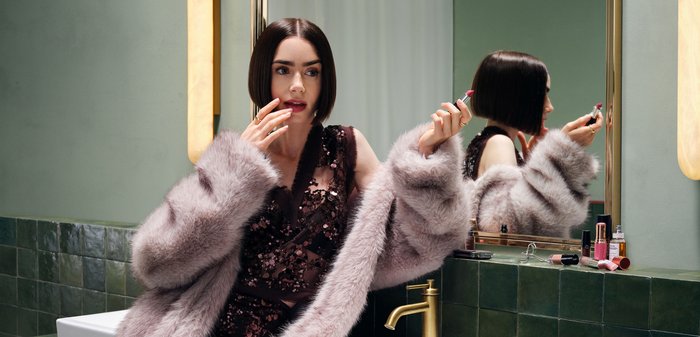 Vrouw in een bontjas brengt rode lippenstift aan terwijl ze in een badkamer spiegel kijkt met cosmetica op het aanrecht.