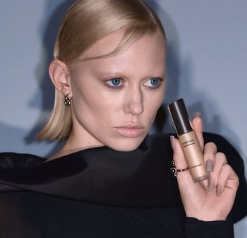 Enlumineur liquide dans un flacon en verre avec une teinte dorée, tenu par une personne portant un haut noir, mettant en valeur un design épuré et une texture lisse.