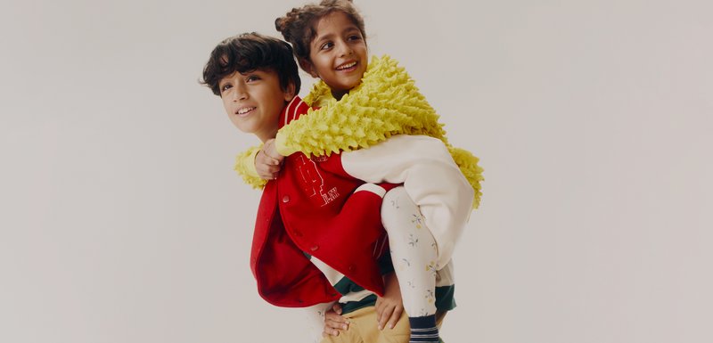 Deux enfants jouent ensemble. L'un porte une veste de varsity rouge ; l'autre est enveloppé dans un châle texturé jaune vif. Tous deux portent des leggings à motifs.