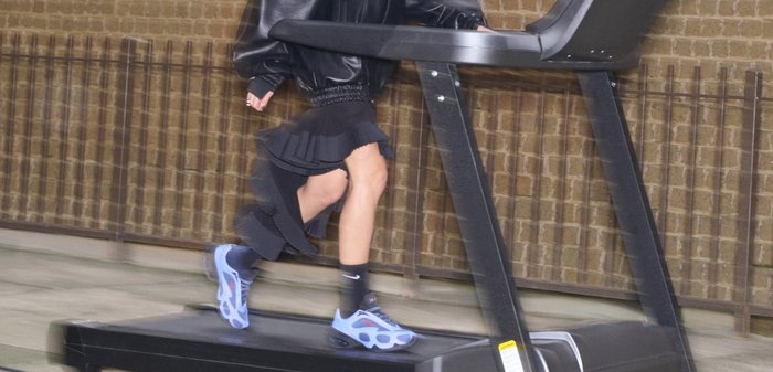 Femme portant une veste noire oversize, une jupe et des baskets bleues, marchant sur un tapis roulant en plein air devant un mur texturé.