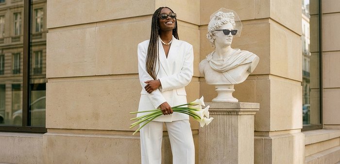Femme en costume blanc tenant des callas blanches, souriante à côté d'un buste classique portant des lunettes de soleil sur un piédestal en pierre en plein air.