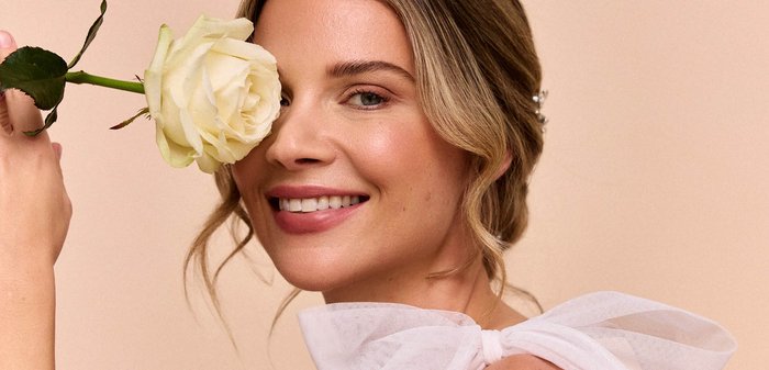 Portrait einer Frau, die eine hellfarbige Rose nahe ihrem Gesicht hält. Sie trägt eine weiße, transparente Schleife um ihren Hals. Sanfte, natürliche Farbtöne.