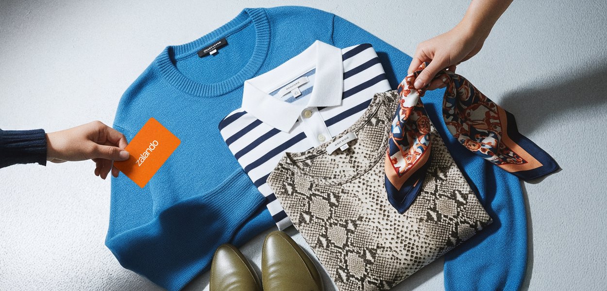Blauwe gebreide trui, gestreepte polo, top met slangenpatroon, kleurrijke sjaal, olijfgroene schoenen en een oranje Zalando-kaart op een oppervlak gerangschikt.